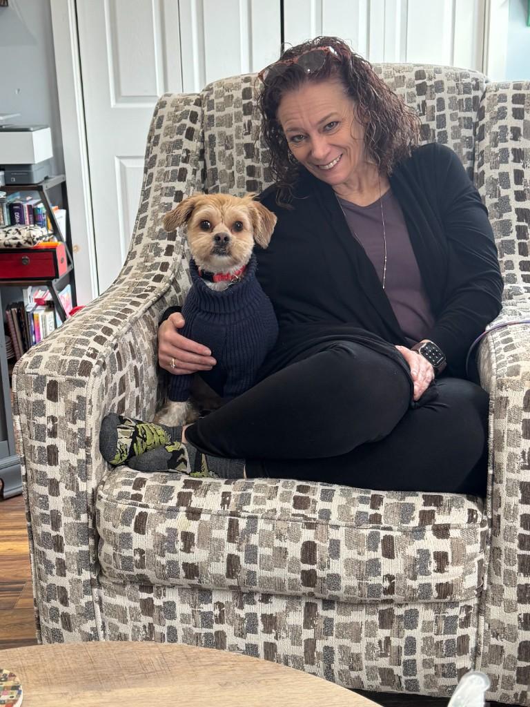 Melissa Gobin Borski seated in a patterned armchair with Roscoe, her small therapy dog in a blue sweater, beside her.