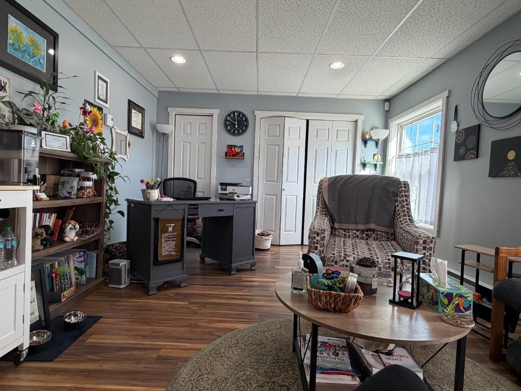 A bright therapy office with a patterned armchair, round wooden coffee table with tissues and an hourglass, bookshelf with plants, and blue-gray walls.